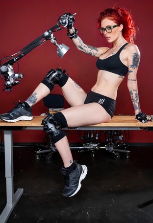 A woman with vibrant red hair, glasses, and tattoos sits on a metal workbench in a high-tech workshop. The metallic d…