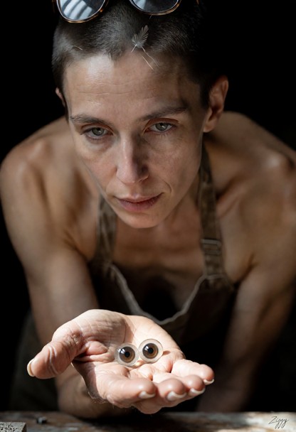 A cinematic photograph in a museum basement workshop, a close shot from the far side of the bench. Her big hand comes…