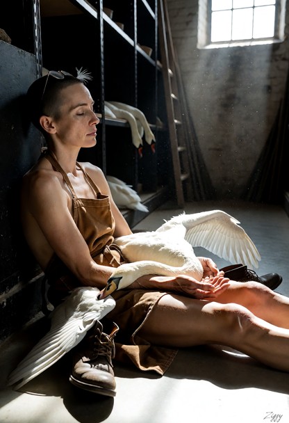 A cinematic photograph in a museum basement workshop, taken from the side at floor level. She sits on the concrete fl…