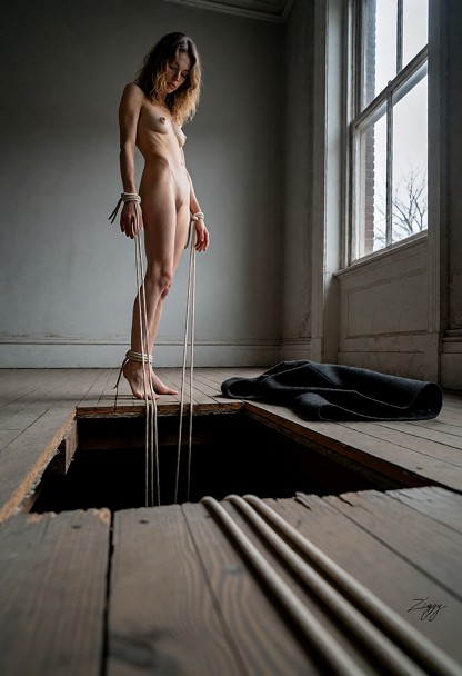 A photograph in the top-floor room of an old building at dawn, empty, one tall window on the right giving flat grey l…