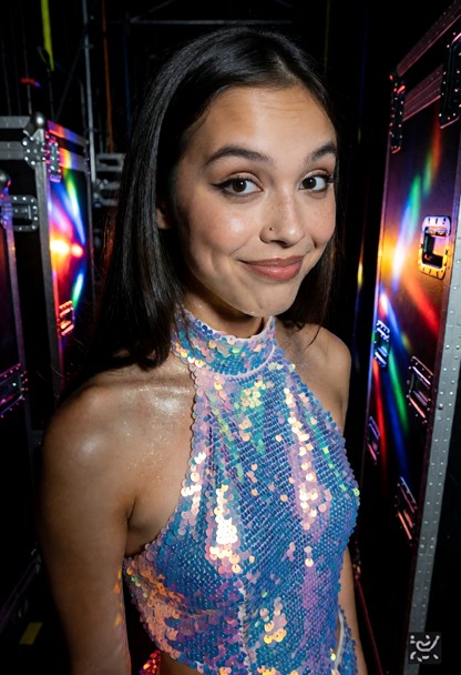Location: dim backstage area filled with shadows and heavy equipment cases, with flickering colorful stage lights ble…