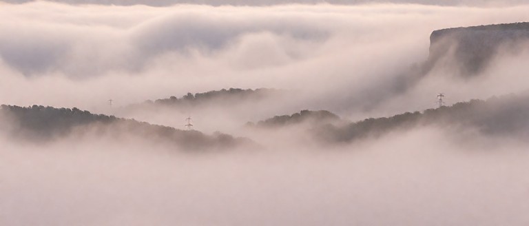 A wide 16:9 photograph dominated by dense, pale fog. Most of the frame contains only soft, opaque mist with a muted d…
