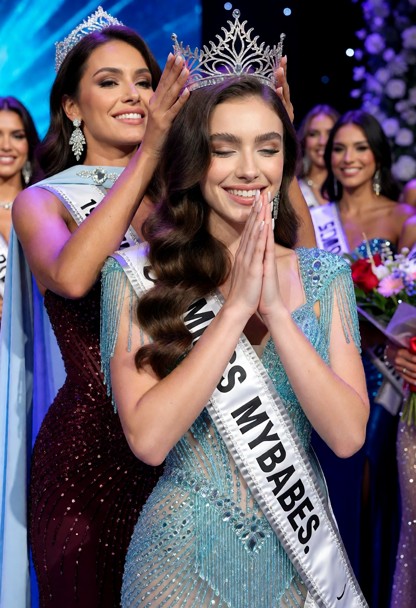 The photograph captures a beauty-pageant crowning ceremony. The winner wears a shimmering turquoise-and-silver gown d…