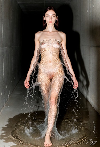 A gown made entirely of clear running water, photographed as a fashion campaign picture in a long bare concrete corri…