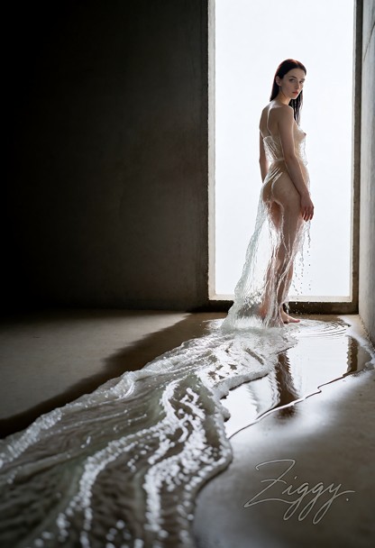 A long bare concrete corridor, the left half of the frame a blank shadowed wall. She stands two thirds of the way dow…